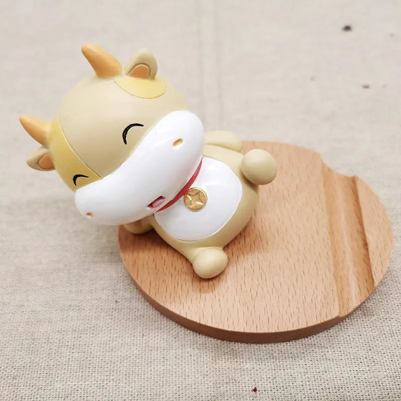 Solid beech wood mobile phone stand with a resin cow figurine and a slot for holding coins on a wooden desk.