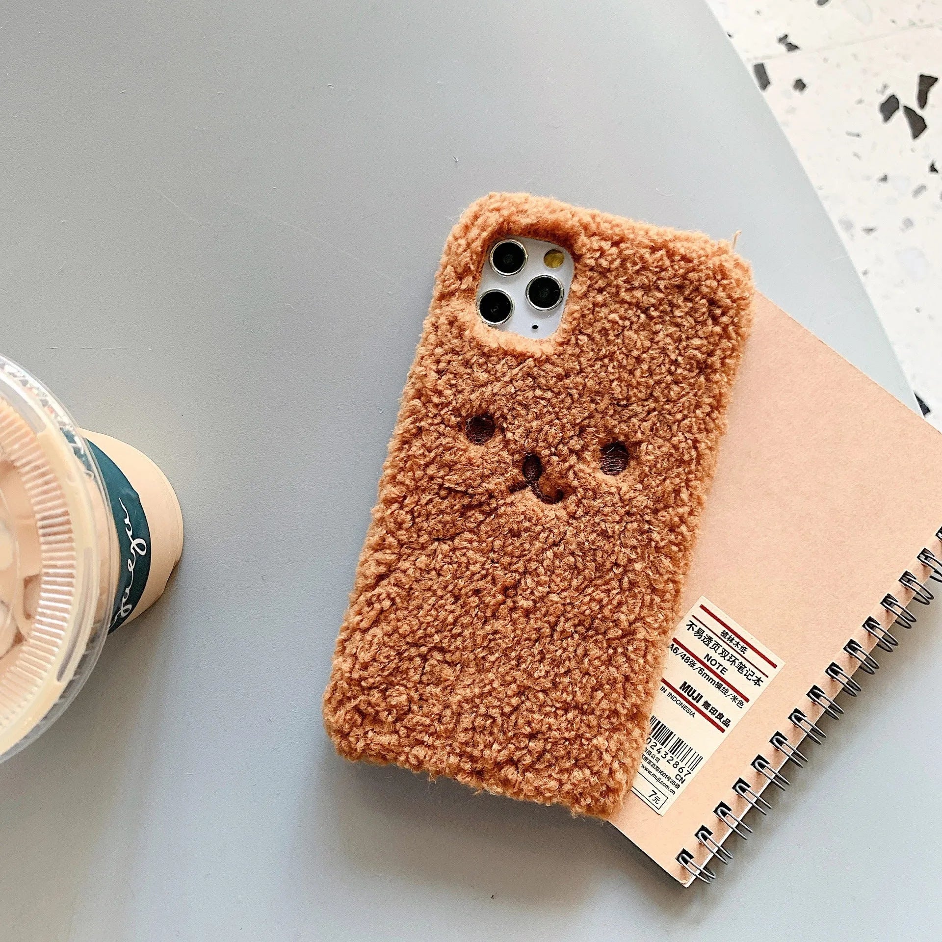 Brown embroidered plush bear face on a clear TPU iPhone case, showing precise cutouts for camera and ports.