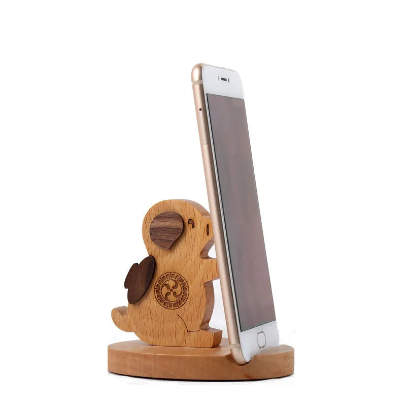 Solid beech wood mobile phone stand with a resin cow figurine and a slot for holding coins on a wooden desk.
