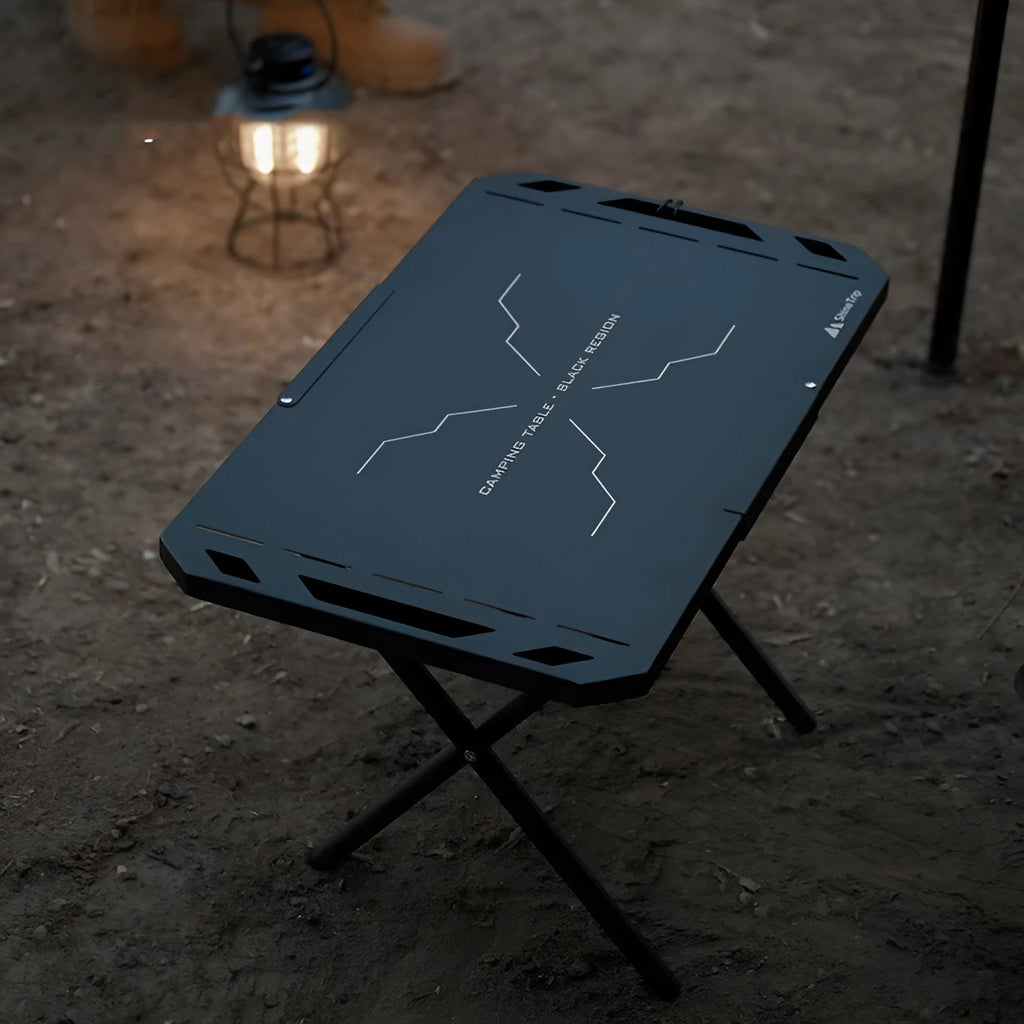 Black folding outdoor camping table with steel frame and storage bag on a campsite