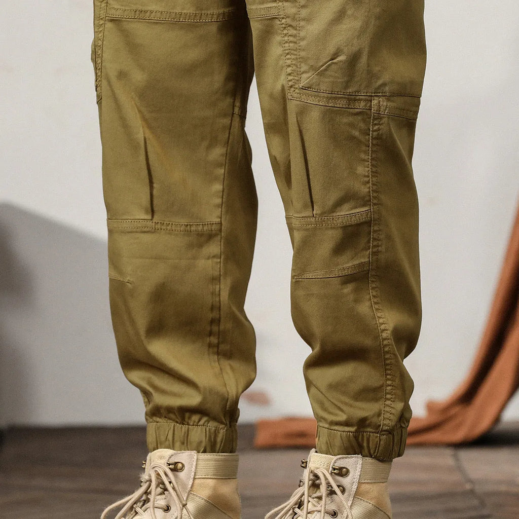 Man wearing khaki coloured cotton overalls with tapered legs and elasticated ankle bands.