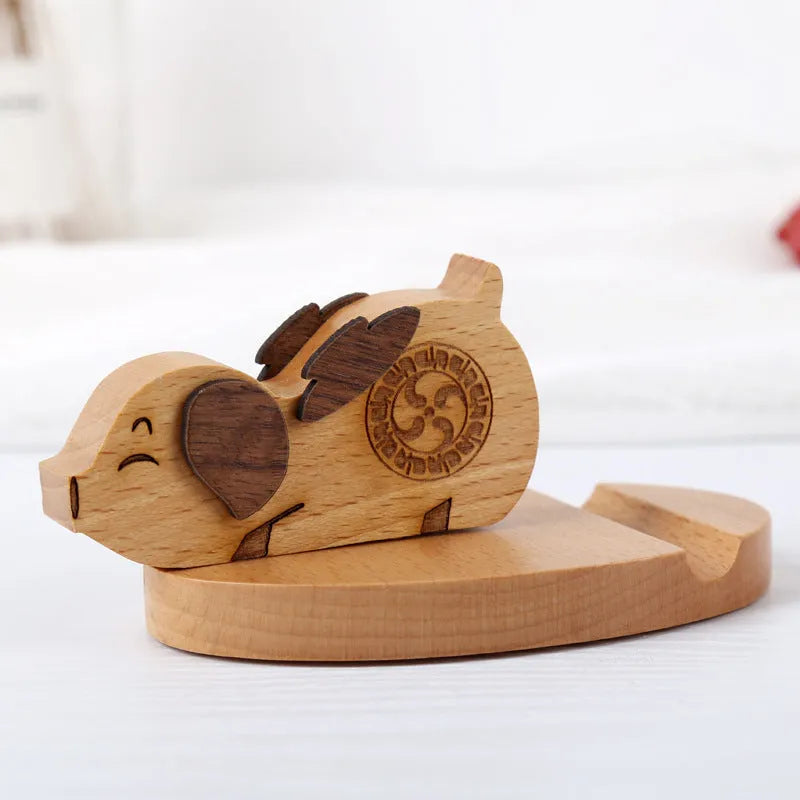 Solid beech wood mobile phone stand with a resin cow figurine and a slot for holding coins on a wooden desk.