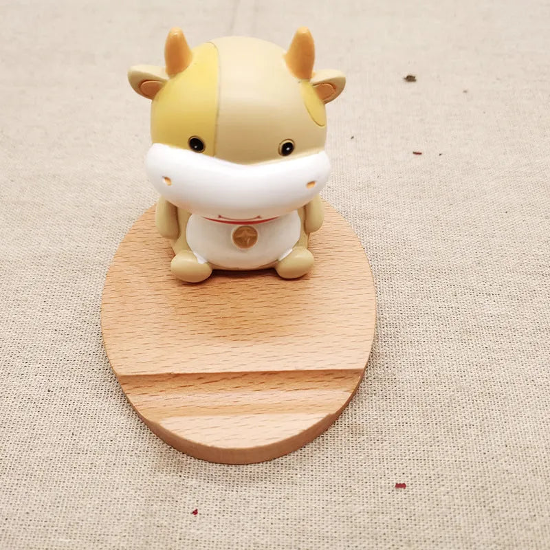 Solid beech wood mobile phone stand with a resin cow figurine and a slot for holding coins on a wooden desk.