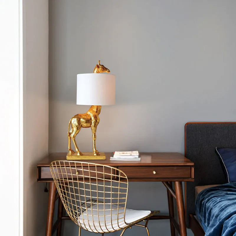 White or gold giraffe-shaped table lamp with cloth shade on a bedside table.