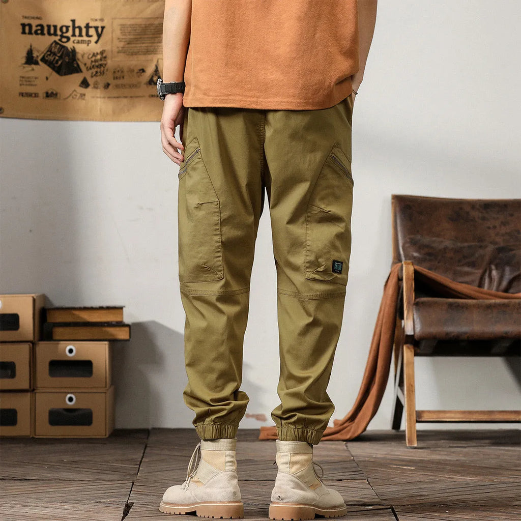 Man wearing khaki coloured cotton overalls with tapered legs and elasticated ankle bands.