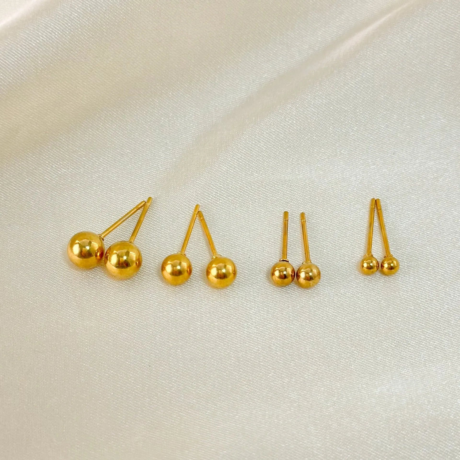 Close-up of minimalist round ball stud earrings in gold and silver finishes on a neutral background.