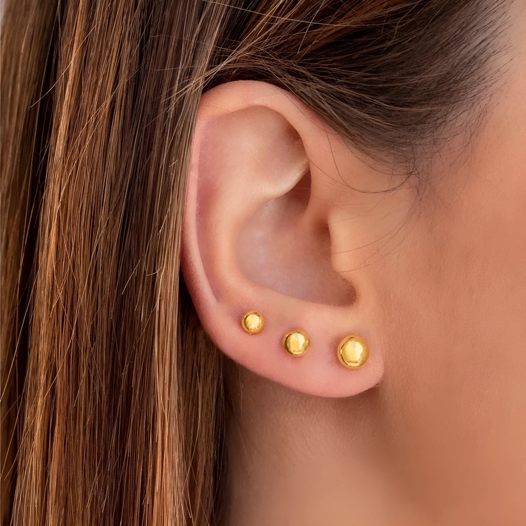 Close-up of minimalist round ball stud earrings in gold and silver finishes on a neutral background.