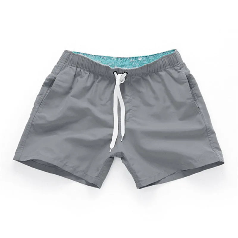 Men's straight-leg polyester surf shorts on a beach, showing colour options like black, fluorescent yellow, and navy blue.