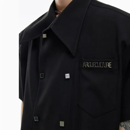 A man wearing a loose-fitting black short-sleeved casual shirt with spliced detailing and metal buttons.