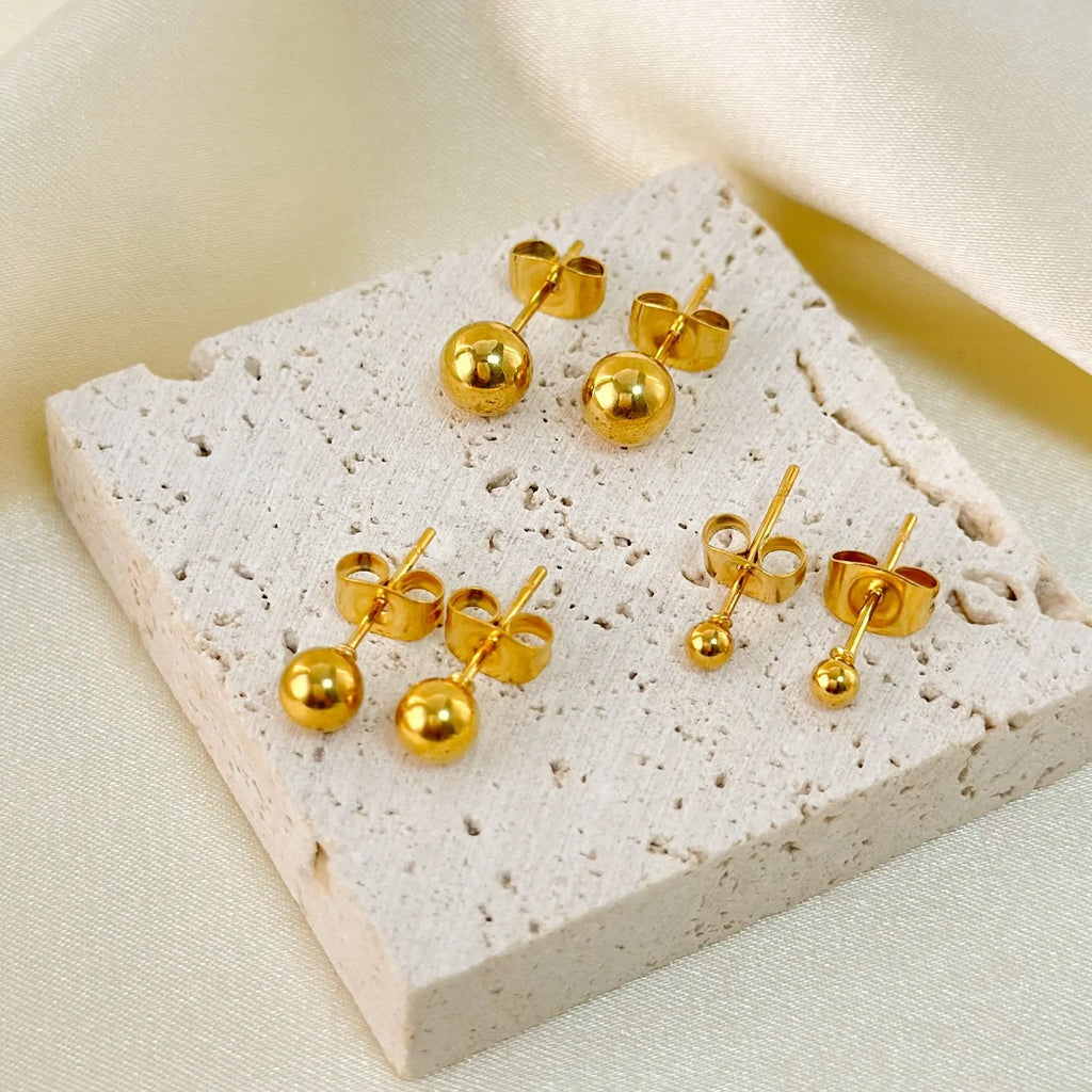 Close-up of minimalist round ball stud earrings in gold and silver finishes on a neutral background.