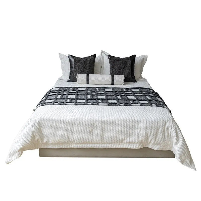 Grey and white plaid bed blanket draped over a dark grey duvet with a matching pillowcase and round leather-look cushion.