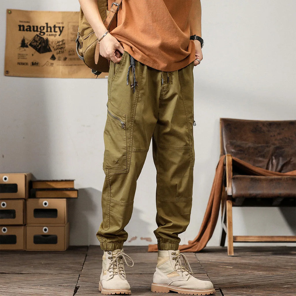 Man wearing khaki coloured cotton overalls with tapered legs and elasticated ankle bands.