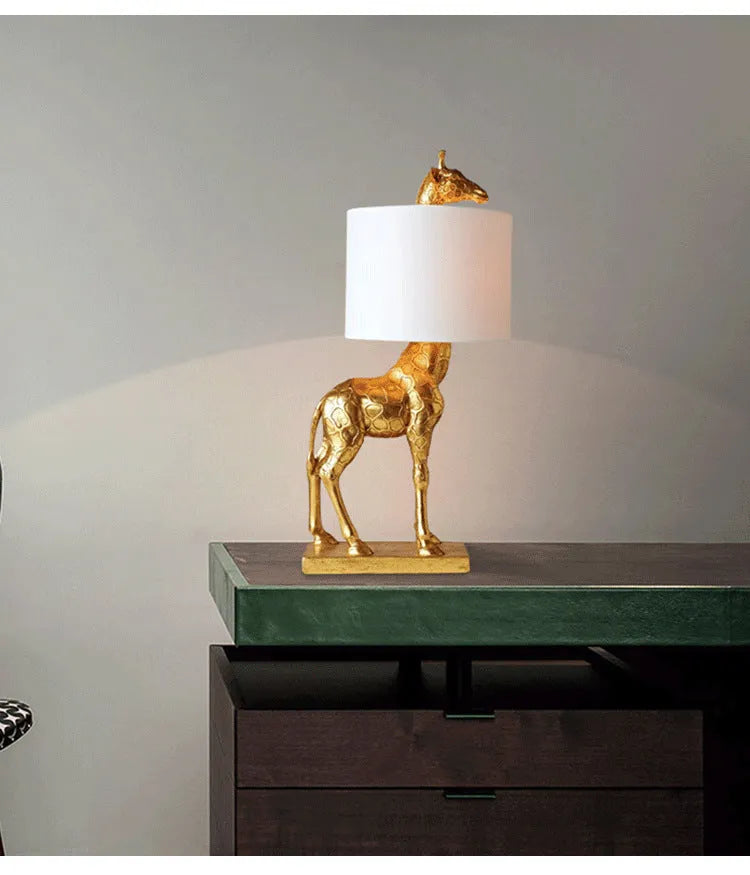 White or gold giraffe-shaped table lamp with cloth shade on a bedside table.