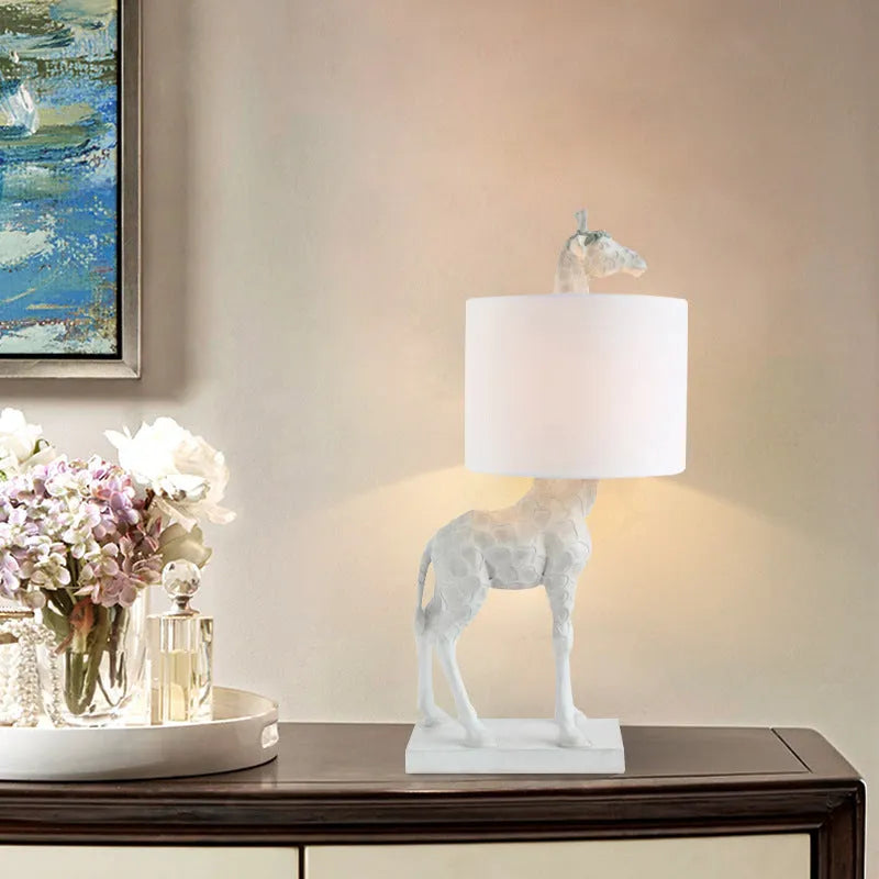 White or gold giraffe-shaped table lamp with cloth shade on a bedside table.