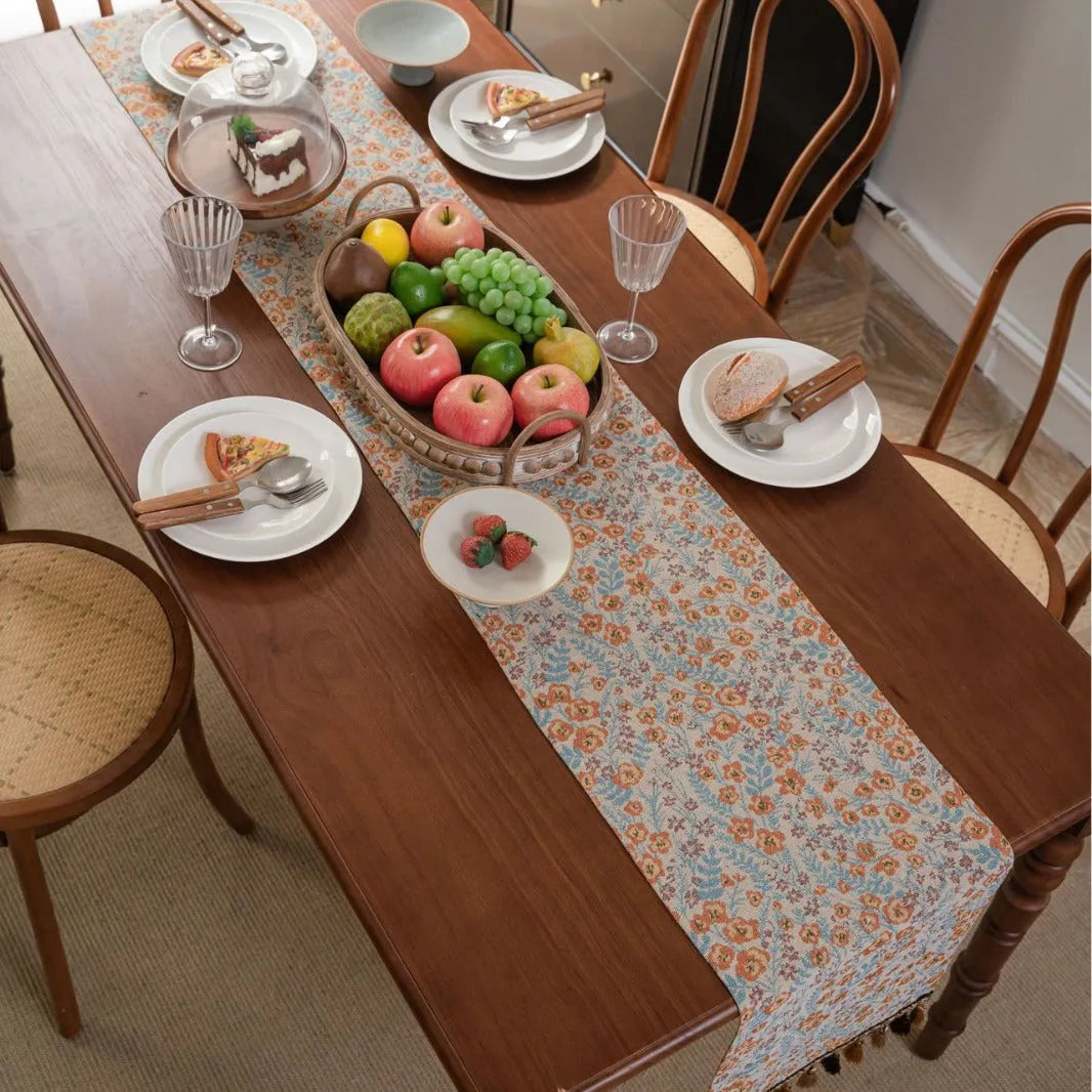 Beige jacquard table runner with flower pattern and long tassels on a wooden dining table.