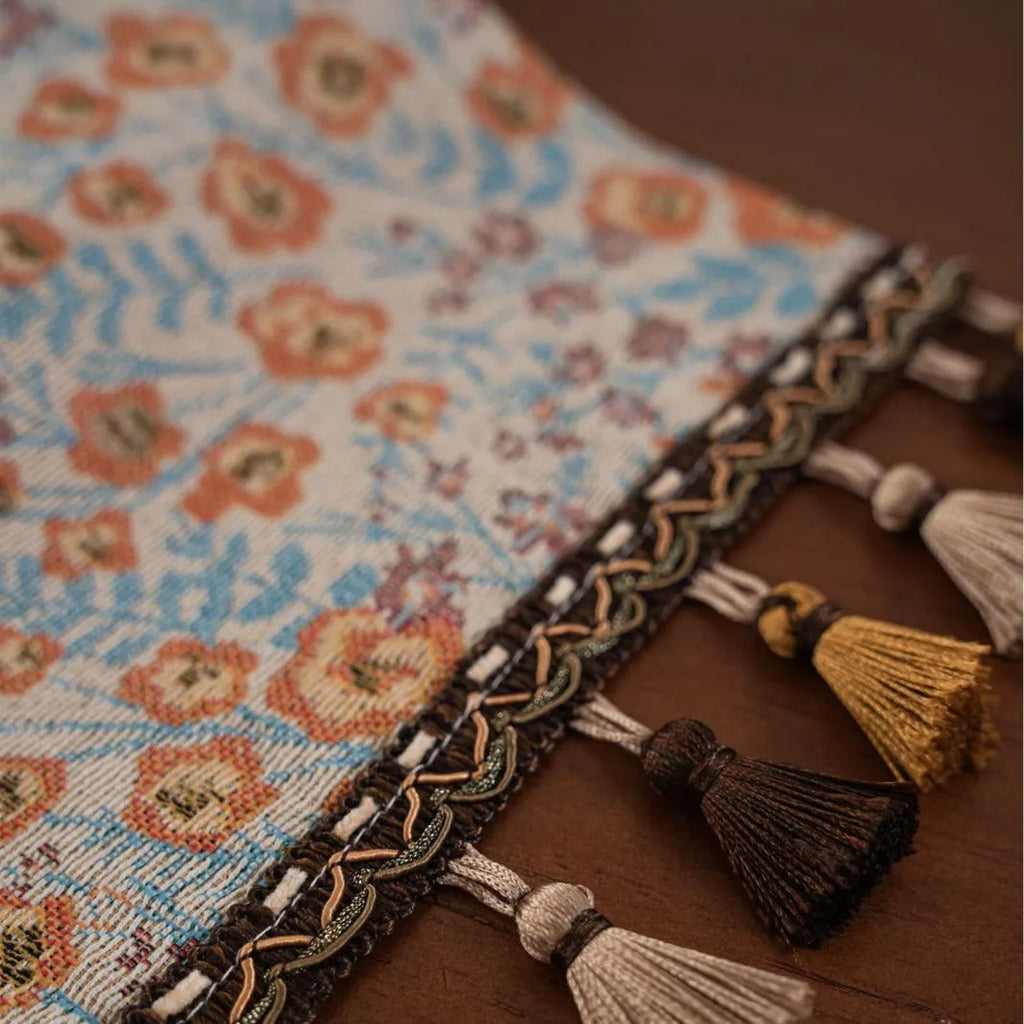 Beige jacquard table runner with flower pattern and long tassels on a wooden dining table.