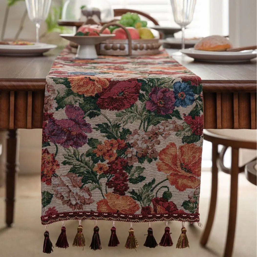 Beige jacquard table runner with flower pattern and long tassels on a wooden dining table.