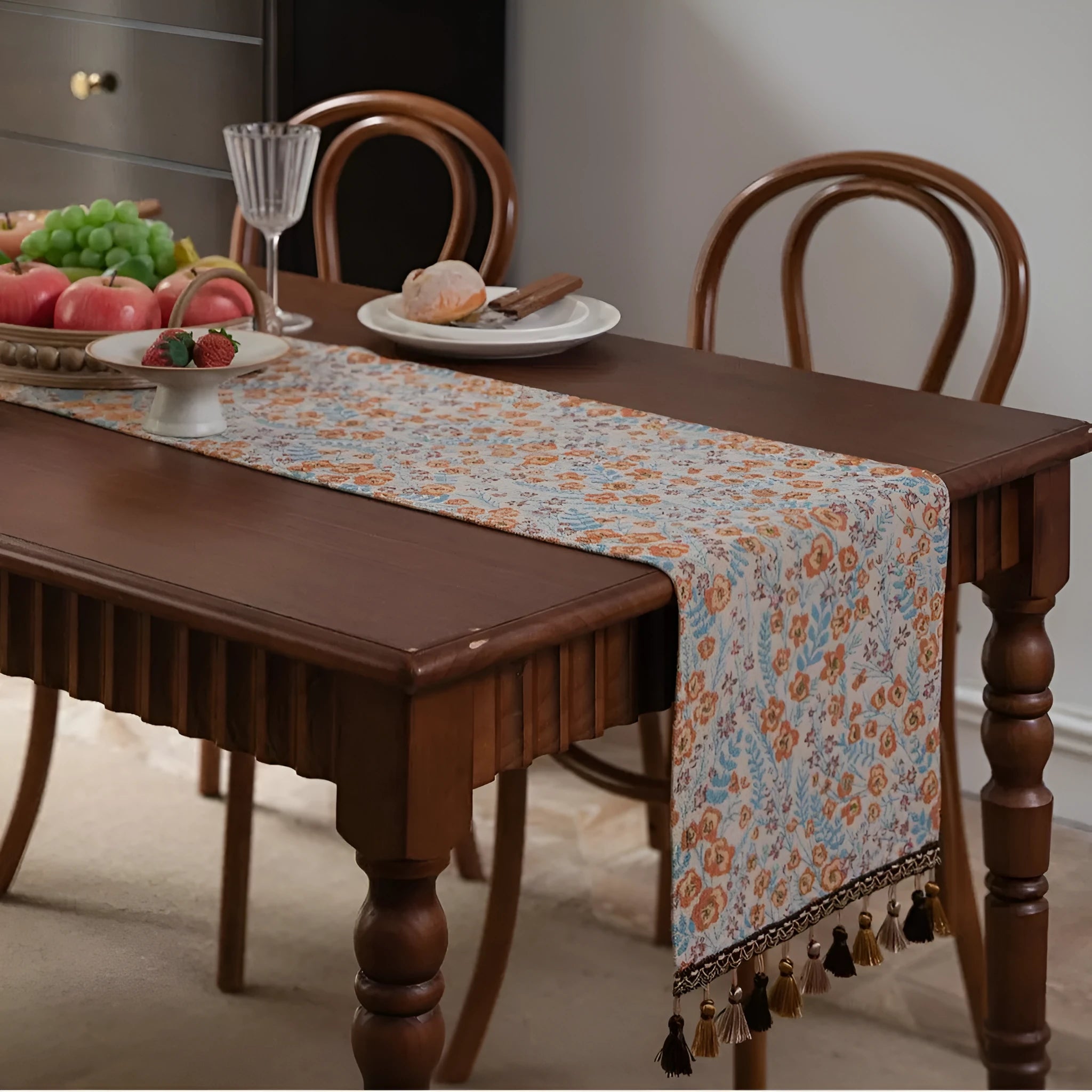 Beige jacquard table runner with flower pattern and long tassels on a wooden dining table.