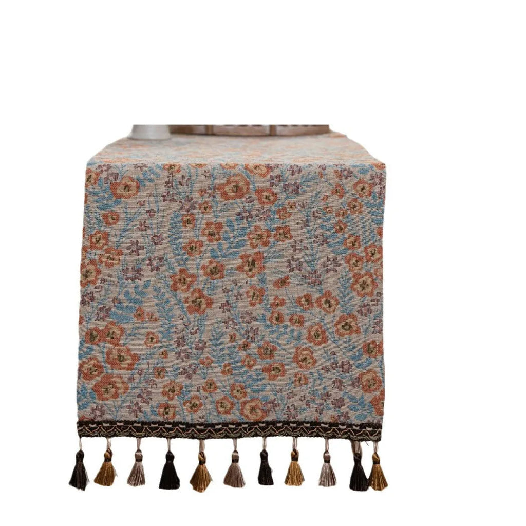 Beige jacquard table runner with flower pattern and long tassels on a wooden dining table.