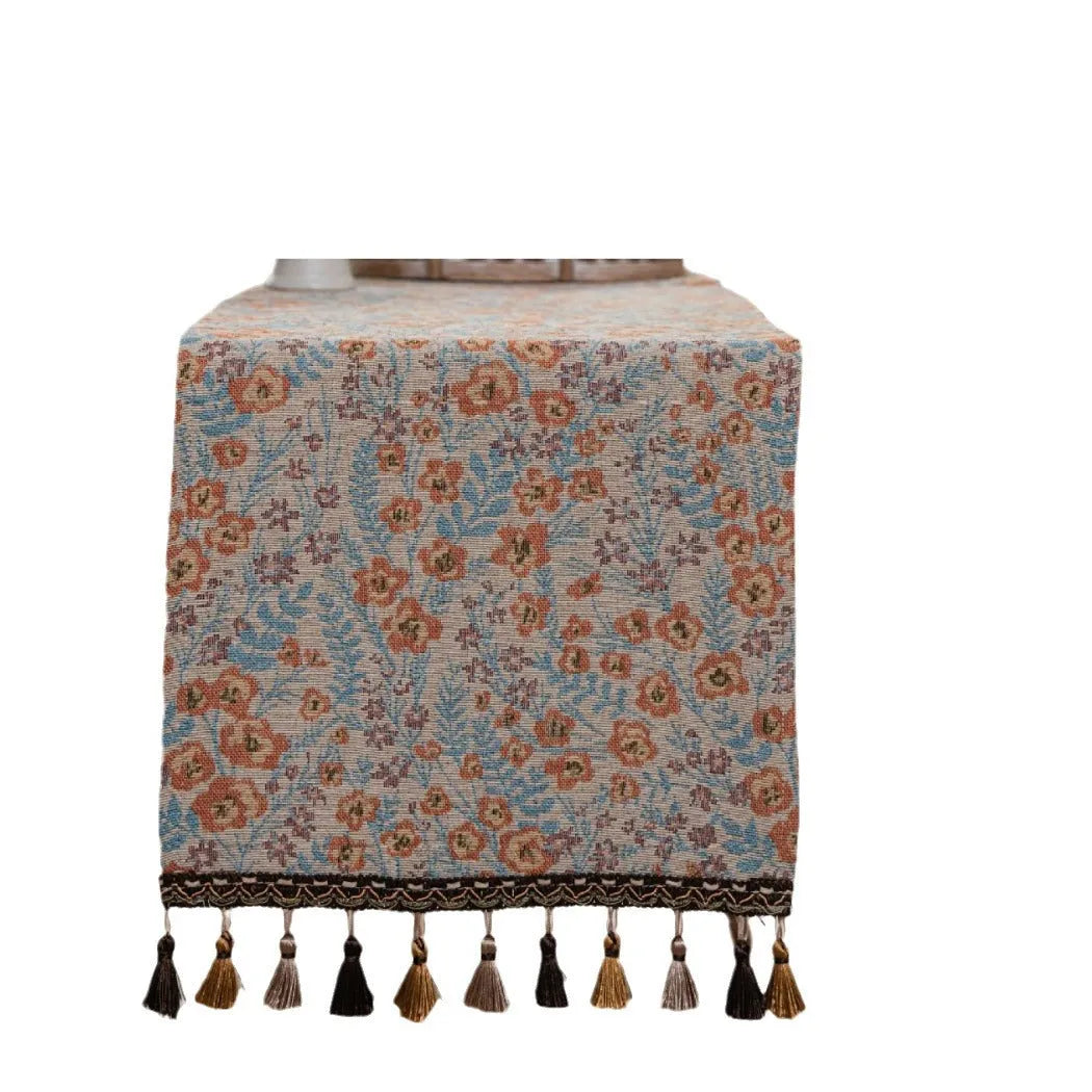 Beige jacquard table runner with flower pattern and long tassels on a wooden dining table.