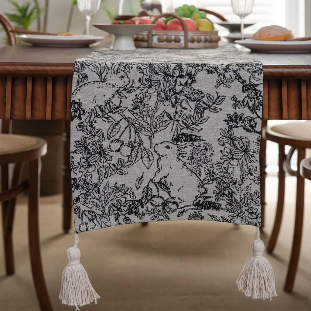 Beige jacquard table runner with flower pattern and long tassels on a wooden dining table.