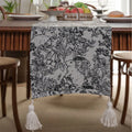 Beige jacquard table runner with flower pattern and long tassels on a wooden dining table.