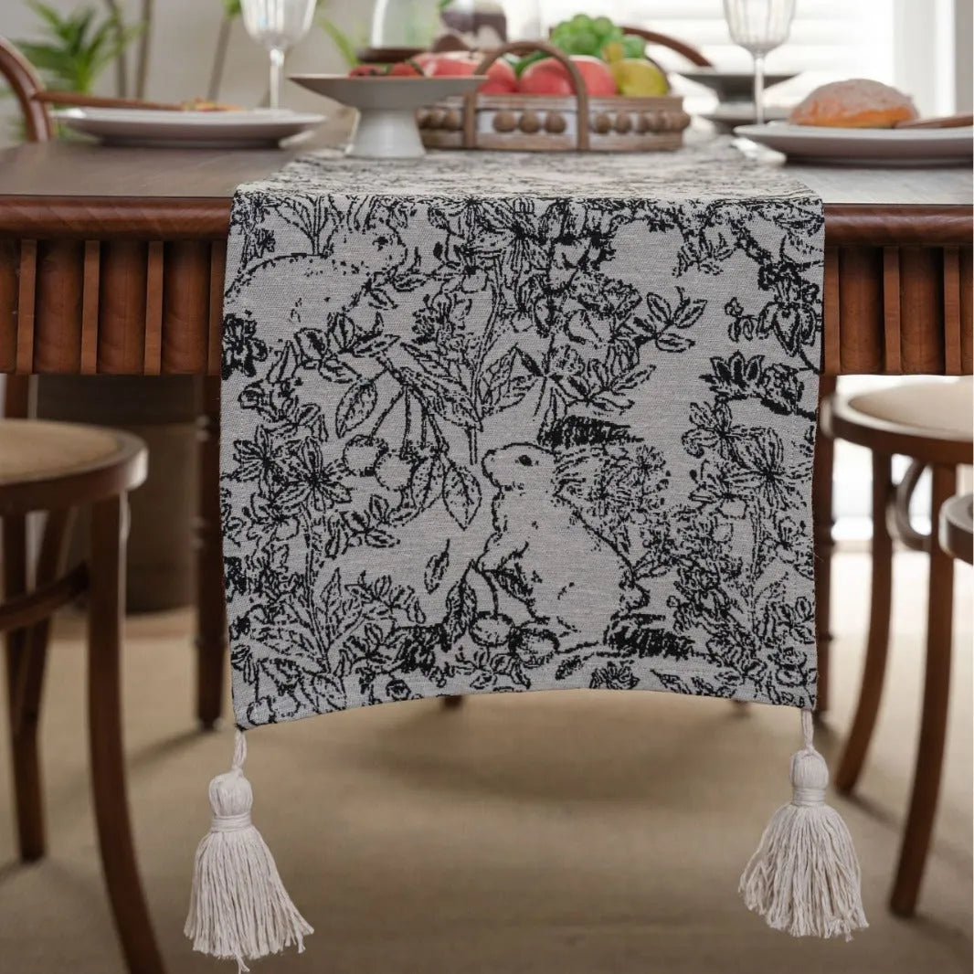 Beige jacquard table runner with flower pattern and long tassels on a wooden dining table.
