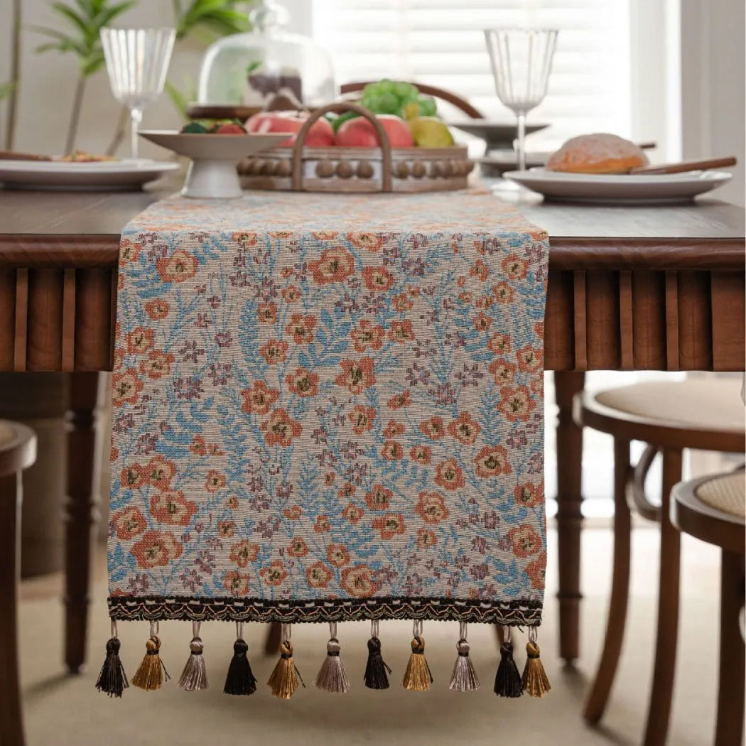 Beige jacquard table runner with flower pattern and long tassels on a wooden dining table.