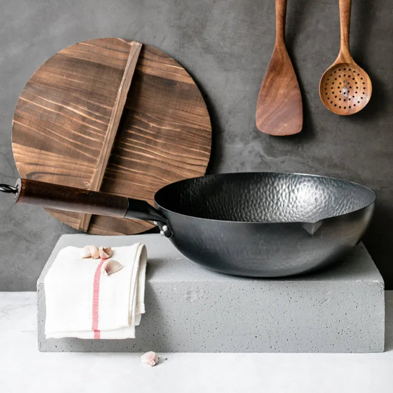 Black handmade traditional iron wok with a long wooden handle, shown with a glass lid.