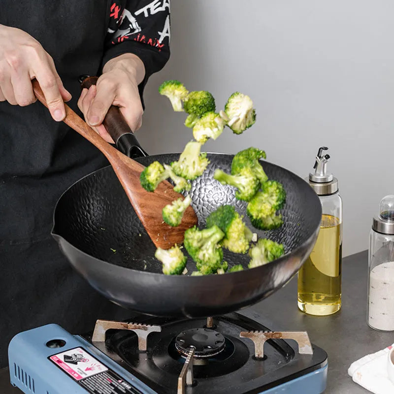 Black handmade traditional iron wok with a long wooden handle, shown with a glass lid.