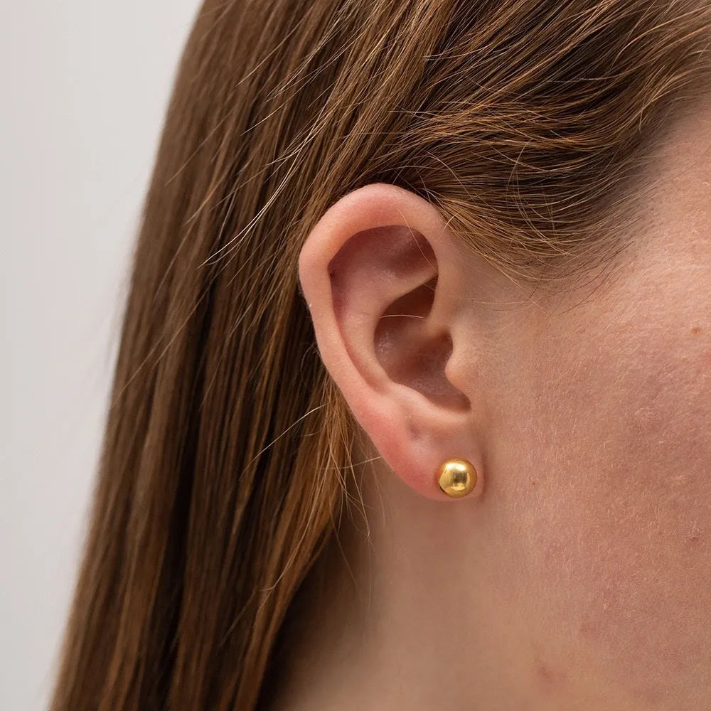 Close-up of minimalist round ball stud earrings in gold and silver finishes on a neutral background.